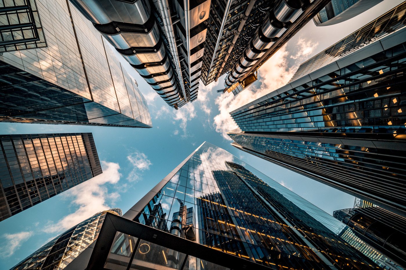 Skyscrapers with glass and metal façades rise toward a blue sky, photographed from ground level looking up.