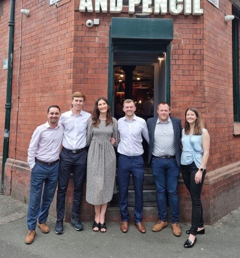 Six adults stand in a row outside a red brick building with a large 'THE PEN AND PENCIL' sign, posing for a group photo.