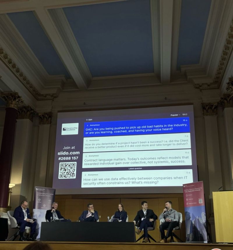 Panel of six speakers seated on a stage in a grand hall, with a large projected screen showing a Slido Q&A chat and branding behind them.